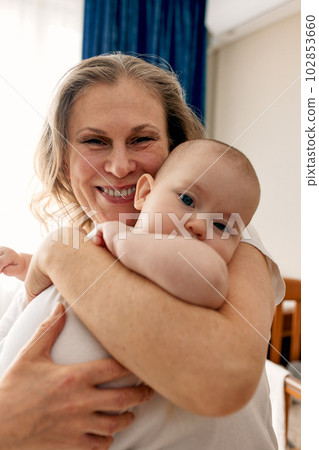 Loving grandmother carries her little granddaughter at home. Bright portrait of a happy grandmother holding her granddaughter in her arms. Grandmother hugs her little granddaughter. 102853660