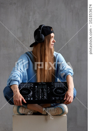 Studio portrait of a young pretty long-haired DJ girl in a blue sweater, loose jeans and a black baseball cap posing while sitting with a DJ console in her hands. Music, fun and creativity. Studio portrait of a young pretty long-haired DJ girl in a blue sweater, loose jeans and a black baseball cap posing while sitting with a DJ console in her hands. Music, fun and creativity. 102853674