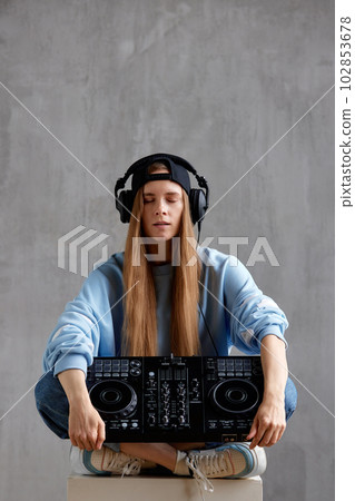 Studio portrait of a young pretty long-haired DJ girl in a blue sweater, loose jeans and a black baseball cap posing while sitting with a DJ console in her hands. Music, fun and creativity. 102853678