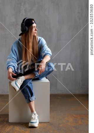 Studio portrait of a young pretty long-haired DJ girl in a blue sweater, loose jeans and a black baseball cap posing while sitting with a DJ console in her hands. Music, fun and creativity. 102853680