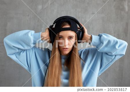 A young pretty long-haired girl in a blue sweater and cap wears DJ headphones on her head and holds them with her hands. Music and beauty. A young pretty long-haired girl in a blue sweater and cap wears DJ headphones on her head and holds them with her hands. Music and beauty. 102853817