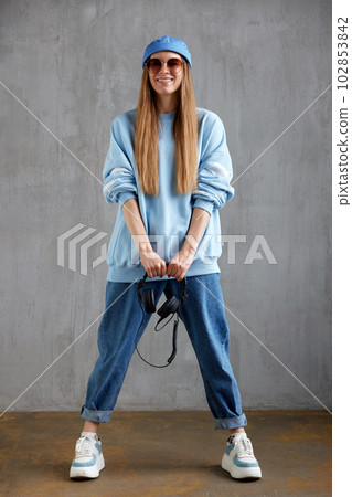 Full length portrait of a young pretty long-haired girl in a blue sweater, cap, jeans and sunglasses holding DJ headphones in her hands. Music, fun and beauty. Full length portrait of a young pretty long-haired girl in a blue sweater, cap, jeans and sunglasses holding DJ headphones in her hands. Music, fun and beauty. 102853842