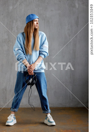 Full length portrait of a young pretty long-haired girl in a blue sweater, cap and jeans holding DJ headphones in her hands. Music, fun and beauty. Full length portrait of a young pretty long-haired girl in a blue sweater, cap and jeans holding DJ headphones in her hands. Music, fun and beauty. 102853849