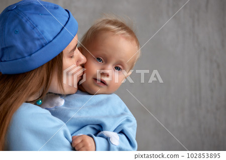 Close-up portrait of a happy young mother and baby infant in her arms. Mom hugs her cute baby. Baby and mother in the same blue clothes. Family love and harmony. 102853895