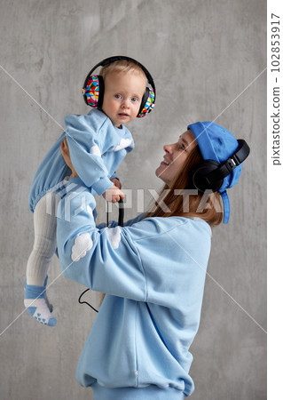 A happy young mother hugs her smiling baby and lifts him up in her arms. Baby and mom in blue clothes and DJ headphones on their heads. 102853917