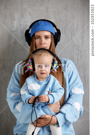 Portrait of a happy young mother and baby infant in her arms. Mom hugs her cute baby. Baby and mother in blue clothes and DJ headphones on their heads. 102853923