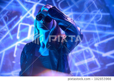 Night life. Urban fashion. Portrait of young beautiful girl in leather jacket posing over blue background with abstract neon elements. 102853966