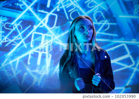 Portrait of young beautiful girl in leather jacket posing, looking away over blue background with abstract neon elements 102853990