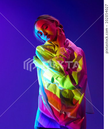 Portrait of young tender girl with multicolored neon filter reflection on body, posing against purple background 102854027