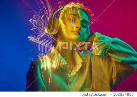 Portrait of young girl listening to music in headphones, posing over gradient blue pink background with mixed neon lights effect 102854095