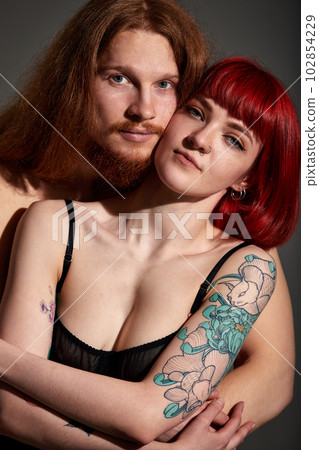 Studio portrait of a sexy half-naked couple in love. A handsome man with long hair and a beard gently hugs his woman with red hair, a piercing and a tattoo. Love, passion and tenderness. Studio portrait of a sexy half-naked couple in love. A handsome man with long hair and a beard gently hugs his woman with red hair, a piercing and a tattoo. Love, passion and tenderness. 102854229
