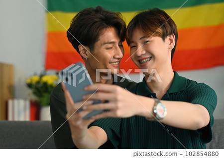 Romantic young gay couple taking a selfie with smartphone in living room rainbow flag in background. LGBT and love concept Romantic young gay couple taking a selfie with smartphone in living room rainbow flag in background. LGBT and love concept 102854880