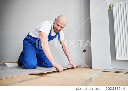 Male construction worker laying laminate wooden plank on floor underlayment. Man in work overalls kneeling down by the wall and installing laminate flooring in living room. Home renovation concept. Male construction worker laying laminate wooden plank on floor underlayment. Man in work overalls kneeling down by the wall and installing laminate flooring in living room. Home renovation concept. 102855644