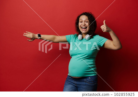 Beautiful pregnant woman with wireless headphones, showing at camera her smart wrist watch, checking contractions and baby kicks on new mobile application for pregnancy, isolated on red background Beautiful pregnant woman with wireless headphones, showing at camera her smart wrist watch, checking contractions and baby kicks on new mobile application for pregnancy, isolated on red background 102855925