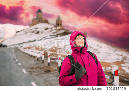 Stepantsminda, Georgia. Man Tourist Backpacker Traveler Photographer In Pink Jacket Posing Near Holy Trinity Church - Tsminda Sameba. Amazing Georgian Landmark In Early Winter. Bold Bright Sky 102855929