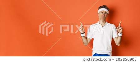 Portrait of skeptical adult sportsman in headband and t-shirt, complaining and pointing fingers up, looking disappointed, standing over orange background Portrait of skeptical adult sportsman in headband and t-shirt, complaining and pointing fingers up, looking disappointed, standing over orange background 102856905