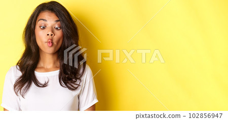 Close-up of funny african-american woman, making faces, squinting and pouting, fooling around over yellow background Close-up of funny african-american woman, making faces, squinting and pouting, fooling around over yellow background 102856947