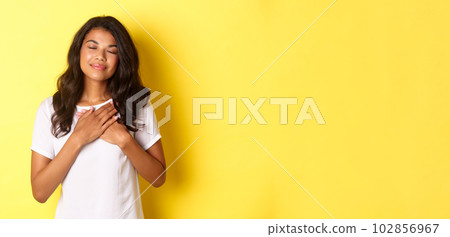 Portrait of romantic african-american woman in white t-shirt, close eyes and hold hands on heart, feeling nostalgic, thinking about someone or having beautiful memory, yellow background 102856967