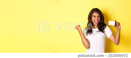 Portrait of cheerful african-american female model, showing credit card and shouting for joy, happy about shopping, standing over yellow background Portrait of cheerful african-american female model, showing credit card and shouting for joy, happy about shopping, standing over yellow background 102856972