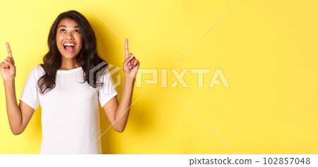 Portrait of amazed, happy african-american girl, smiling and pointing fingers up, standing over yellow background Portrait of amazed, happy african-american girl, smiling and pointing fingers up, standing over yellow background 102857048