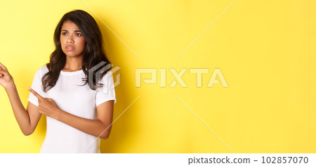 Portrait of doubtful and anxious african-american woman, frowning and pointing fingers at upper left corner, showing something disturbing, standing over yellow background Portrait of doubtful and anxious african-american woman, frowning and pointing fingers at upper left corner, showing something disturbing, standing over yellow background 102857070
