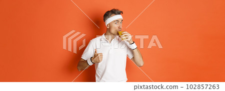 Concept of workout, gym and lifestyle. Portrait of handsome middle-aged fitness guy, showing thumbs-up and drinking juice, standing over orange background 102857263