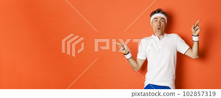 Concept of sport, fitness and lifestyle. Portrait of amazed and excited middle-aged male athlete, pointing fingers sideways, showing two variants, standing over orange background 102857319