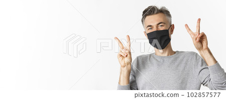 Concept of covid-19, social distancing and quarantine. Close-up of happy and cute middle-aged guy in black medical mask, showing peace signs and smiling, standing over white background Concept of covid-19, social distancing and quarantine. Close-up of happy and cute middle-aged guy in black medical mask, showing peace signs and smiling, standing over white background 102857577