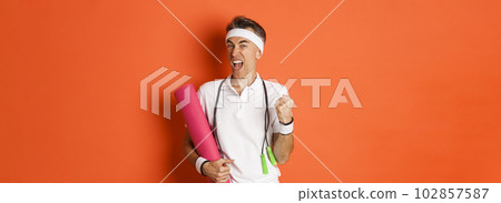Concept of workout, gym and lifestyle. Image of motivated and happy middle-aged fitness guy, achieve goal, saying yes and making fist pump, holding yoga mat with skipping rope 102857587