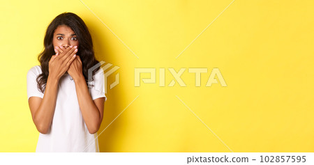 Portrait of timid and scared african-american girl, shut mouth and looking anxious, standing frightened over yellow background 102857595