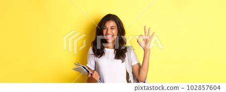 Portrait of confident african-american female student, showing okay sign and winking, approve something good, standing with notebooks and backpack for college, yellow background Portrait of confident african-american female student, showing okay sign and winking, approve something good, standing with notebooks and backpack for college, yellow background 102857604