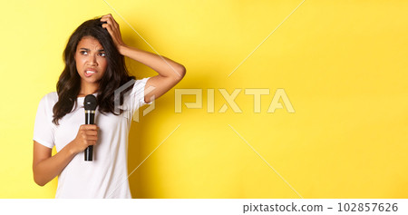 Image of indecisive african-american girl, holding microphone and looking hesitant, standing over yellow background Image of indecisive african-american girl, holding microphone and looking hesitant, standing over yellow background 102857626