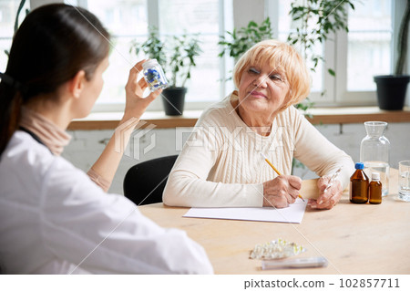 Young woman, doctor, therapist prescribing pill, meds to her senior female patient. Modern hospital, office. Young woman, doctor, therapist prescribing pill, meds to her senior female patient. Modern hospital, office. 102857711