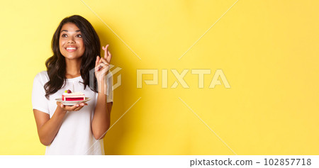 Image of cute and dreamy african-american girl, crossing fingers, holding birthday cake and looking left while making a wish, celebrating b-day over yellow background 102857718