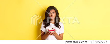 Image of indecisive and sad african-american woman, looking upset, cannot eat cake, standing over yellow background 102857724