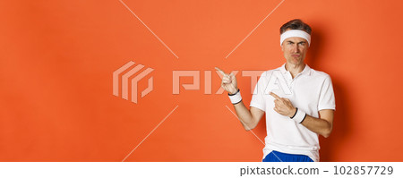 Portrait of disappointed middle-aged man in sportswear, complaining and pointing fingers left at logo, showing something bad, standing over orange background Portrait of disappointed middle-aged man in sportswear, complaining and pointing fingers left at logo, showing something bad, standing over orange background 102857729