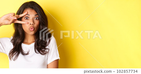 Image of silly and cute teenage african-american girl, showing peace sign and pouting for kiss, standing over yellow background 102857734