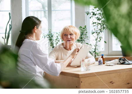 Senior woman visiting doctor, sitting at table in office, having appointment and consultation. Nurse showing information on tablet 102857735