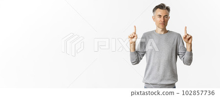 Image of thoughtful and unsure adult man, squinting hesitant while pointing fingers up, making choice, standing over white background 102857736