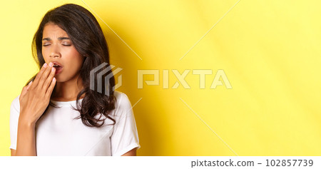 Close-up of tired and sleepy african-american girl, yawning and looking exhausted, standing in white t-shirt over yellow background 102857739