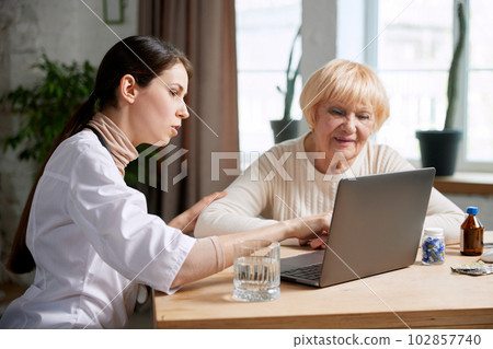 Female doctor sitting at table in office with her senior patient and talking, looking on laptop. Explaining medical results 102857740