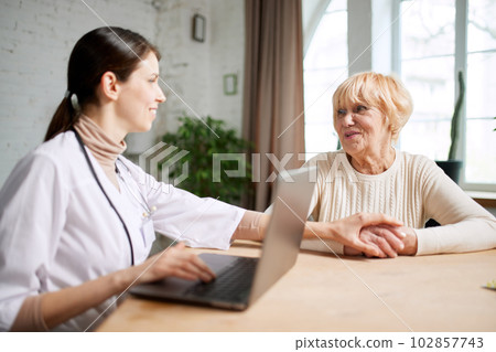 Female doctor sitting at table in office with her senior patient and talking, looking on laptop. Information about health 102857743