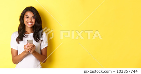 Image of modern african-american girl smiling, using mobile phone, standing over yellow background Image of modern african-american girl smiling, using mobile phone, standing over yellow background 102857744