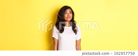 Portrait of hopeful young african-american girl, longing for something, looking at upper left corner dreamy, standing over yellow background 102857750