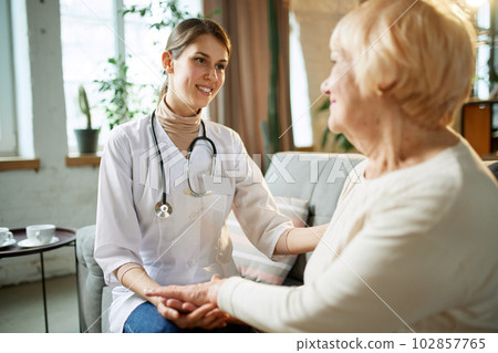 Smiling young woman, doctor giving support and hope to her elderly female patient. Woman holding hand of senior lady at home 102857765