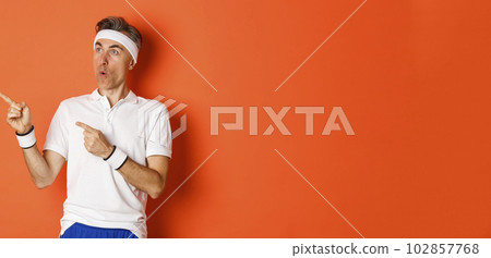 Image of intrigued middle-aged sportsman in activewear, pointing and looking left at advertisement about workout or sports, standing over orange background 102857768