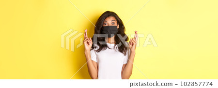 Concept of covid-19, social distancing and lifestyle. Image of excited and hopeful african-american girl, making a wish, wearing face mask and crossing fingers for good luck, over yellow background 102857774