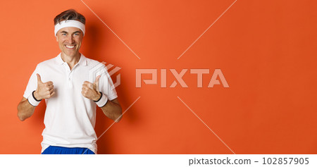 Concept of sport, fitness and lifestyle. Portrait of confident, attractive middle-aged male athlete in workout uniform, showing thumbs-up in approval and smiling, orange background 102857905