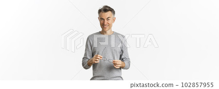 Portrait of cheeky smiling man in grey sweater, holding glasses and looking cunning at camera, standing over white background Portrait of cheeky smiling man in grey sweater, holding glasses and looking cunning at camera, standing over white background 102857955