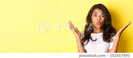 Close-up of surprised african-american girl, raising hands up and saying wow, hear amazing news, standing over yellow background Close-up of surprised african-american girl, raising hands up and saying wow, hear amazing news, standing over yellow background 102857993